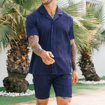 Model wearing blue men's casual button-up shirt and shorts set against tropical backdrop.